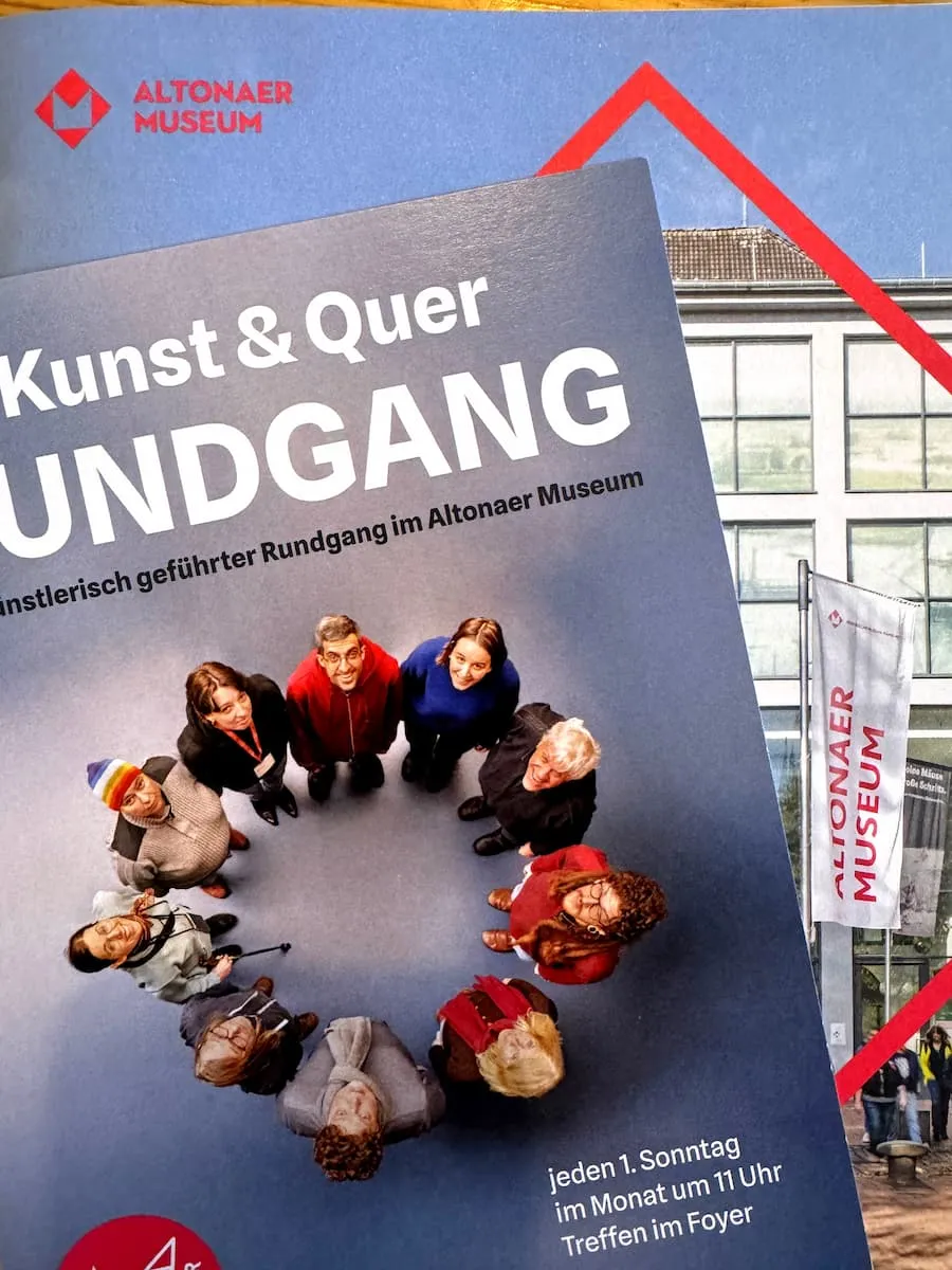 Ein Flyer für den Rundgang „Kunst und Quer“. Darauf 10 Personen im Kreis, von oben fotografiert. Hinter dem Flyer ein Plakat mit dem Aufdruck und Foto vom Altonaer Museum.