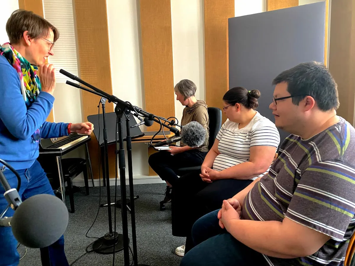 Vier Menschen in einem Tonstudio. Zwei Frauen und ein Mann sitzen vor Mikrofonen, eine Frau steht.