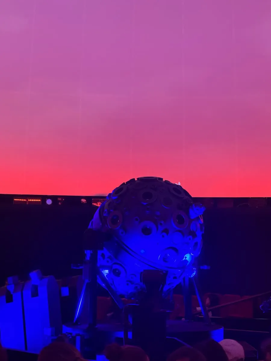 ein großer, runder Sternenprojektor in einem Planetarium. Dahinter ein pink-violetter Himmel.