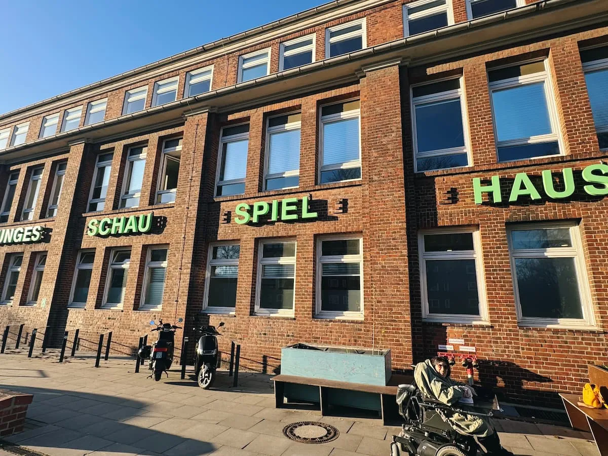 Eine Frau im Rollstuhl steht vor einem roten Backsteingebäude. Auf dem Gebäude steht in großen grünen Buchstaben „Junges Schauspielhaus“