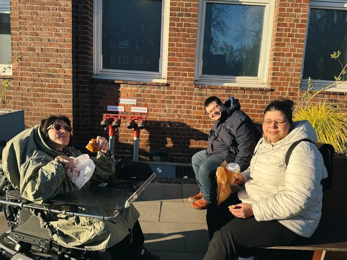 Drei Personen vor einer roten Backsteinwand mit Fenstern. Eine Frau sitzt im Rollstuhl.